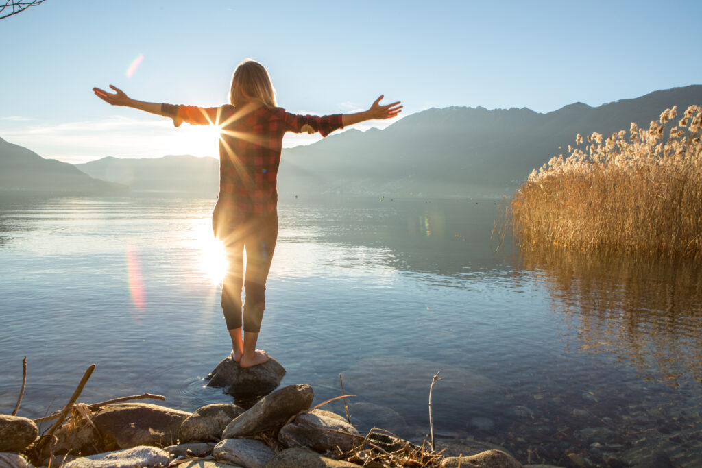 Woman standing on a rock at a peaceful lake with arms open wide, symbolizing the freedom, support, and healing offered through individual therapy using ACT, mindfulness, and trauma-informed care