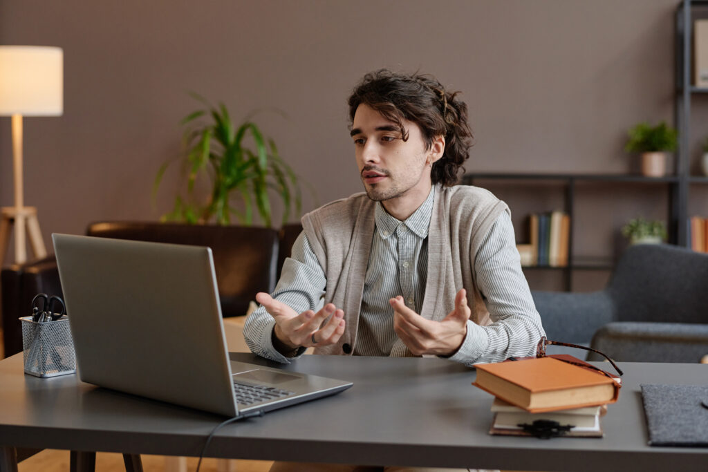 Person engaged in online individual therapy session via laptop, accessing support from home with books and a cozy office background